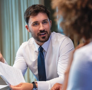 A man in a suit talking with a woman with a paper in his hand.